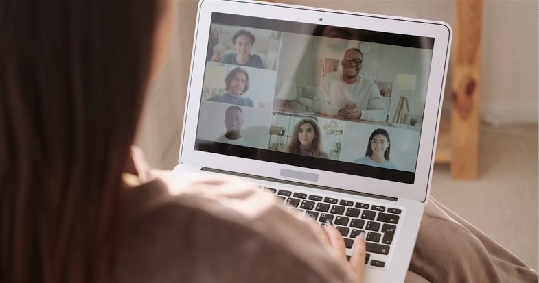 Symbolbild: Eine Person ist von hinten zu sehen, sie schaut auf einen Laptop auf dem mehrere Personen zu sehen sind, die in einem digitalen Meeting miteinander sprechen.