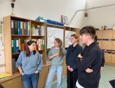 Leipziger Studierende stehem mit einer Lehrkraft der Mittelschule Puntigam im Halbkreis im Gespräch in einem Schulraum mit Bücherschränken im Hintergrund