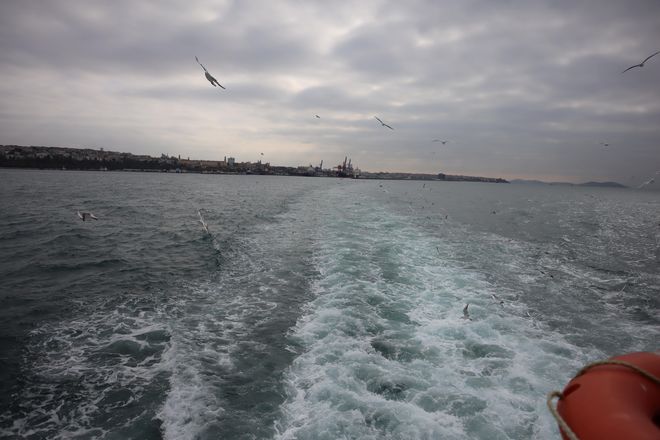 Von dem hinteren Ende der Fähre wird der Bosporus fotografiert. Die Fähre hinterlässt kleine Wellen. Möwen fliegen über dem Wasser. Im Hintergrund ist Istanbul zu sehen.