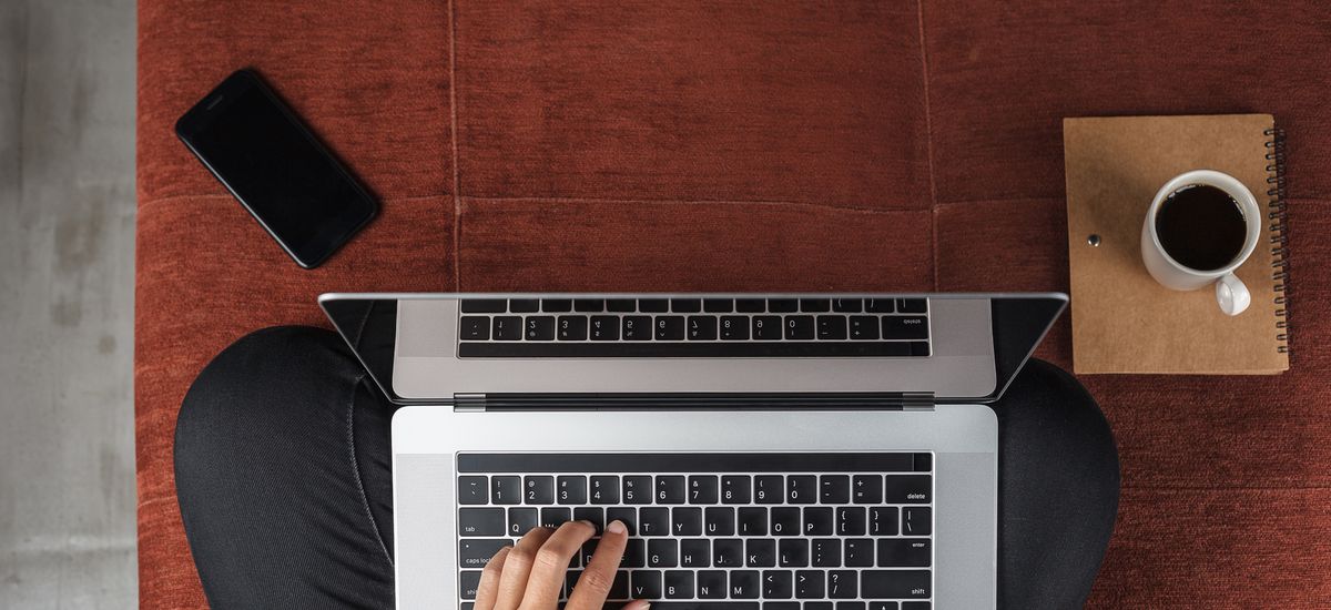 Vorgelperspektive auf einen Menschen, der im Schneidersitz auf dem Fußboden sitzt. Auf dem Schoß steht der Laptop. Die Hände liegen auf der Tastatur und tippen. Der Rest vom Körper ist nicht zu sehen.