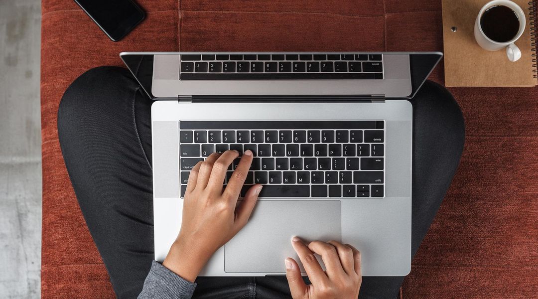 Vorgelperspektive auf einen Menschen, der im Schneidersitz auf dem Fußboden sitzt. Auf dem Schoß steht der Laptop. Die Hände liegen auf der Tastatur und tippen. Der Rest vom Körper ist nicht zu sehen.
