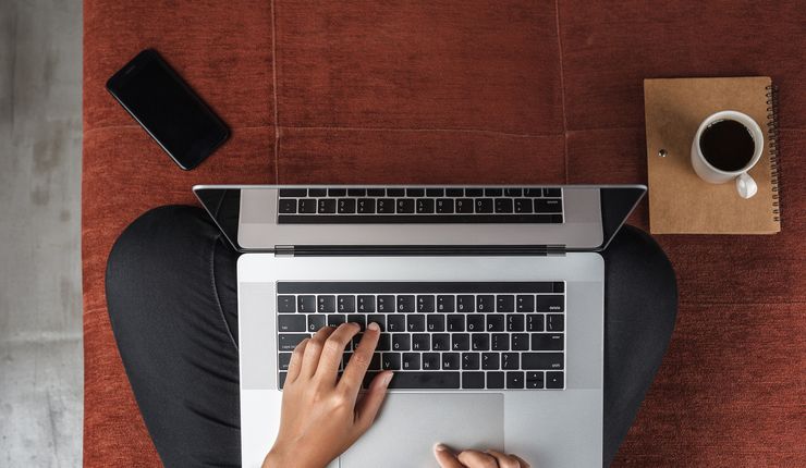 Vorgelperspektive auf einen Menschen, der im Schneidersitz auf dem Fußboden sitzt. Auf dem Schoß steht der Laptop. Die Hände liegen auf der Tastatur und tippen. Der Rest vom Körper ist nicht zu sehen.