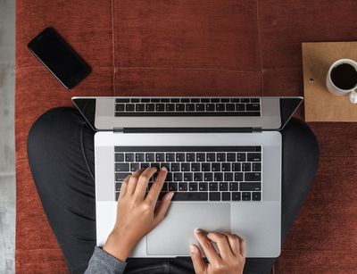 Vorgelperspektive auf einen Menschen, der im Schneidersitz auf dem Fußboden sitzt. Auf dem Schoß steht der Laptop. Die Hände liegen auf der Tastatur und tippen. Der Rest vom Körper ist nicht zu sehen.