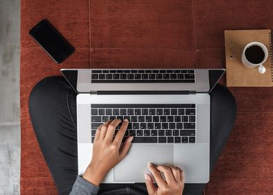 Vorgelperspektive auf einen Menschen, der im Schneidersitz auf dem Fußboden sitzt. Auf dem Schoß steht der Laptop. Die Hände liegen auf der Tastatur und tippen. Der Rest vom Körper ist nicht zu sehen.