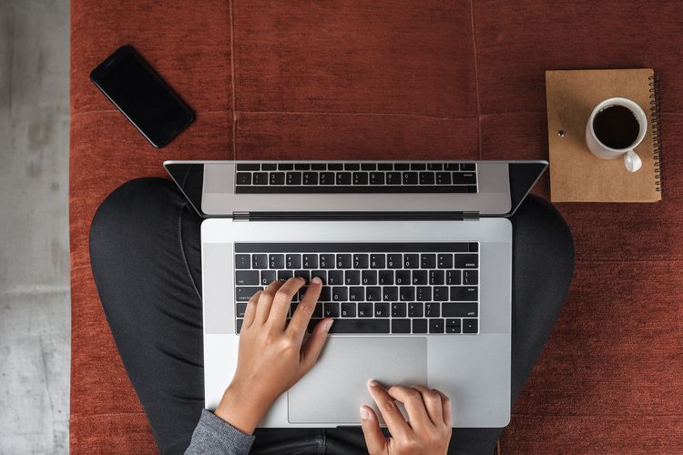 Vorgelperspektive auf einen Menschen, der im Schneidersitz auf dem Fußboden sitzt. Auf dem Schoß steht der Laptop. Die Hände liegen auf der Tastatur und tippen. Der Rest vom Körper ist nicht zu sehen.