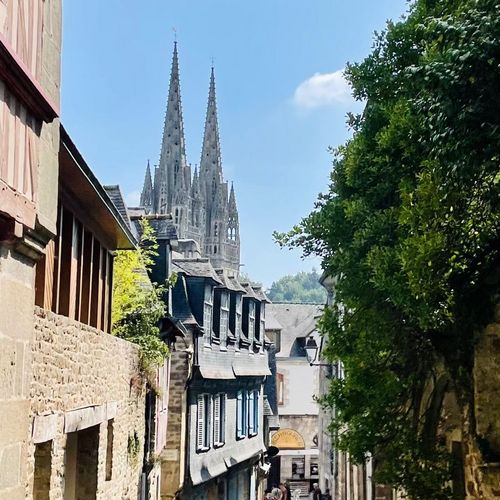 Der Kirchturm der Stadt Quimper ist im linken Bildhintergrund zu sehen. Rechts wird das Bild von Bäumen gesäumt. Links befinden sich Häuser.