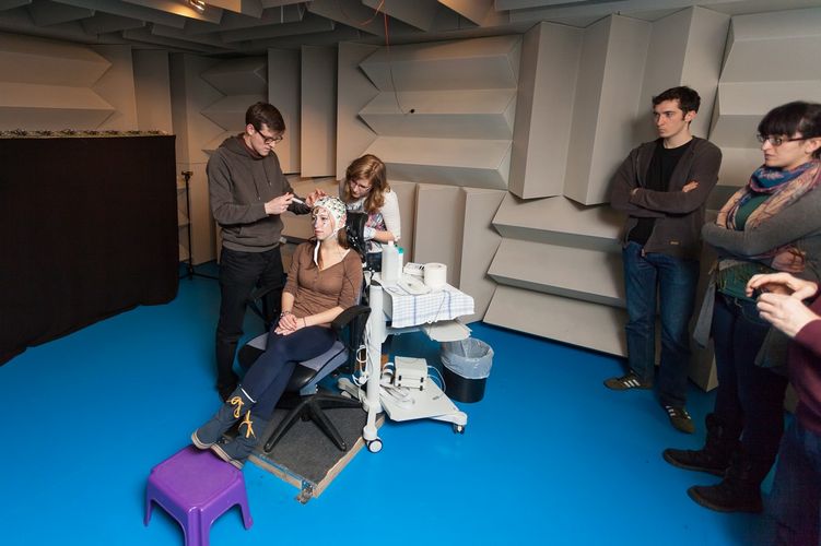 Zwei Studierende sind in einem schallisolierten Raum und führen ein Experiment an einer Probantin durch, die eine Kopfhaube mit Kabeln trägt, Foto: Christian Hüller