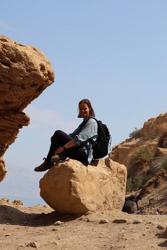 Eine Frau sitzt mit angezogenen Beinen auf einem kleinen Felsen. Neben ihr Ragen größere Felsen empor. Im Hintergrund ist ein See zu erahnen.