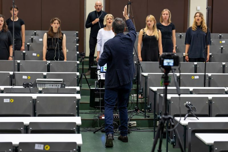 zur Vergrößerungsansicht des Bildes: Farbfoto: Im Vordergrund sind Tische und Stühle eines Seminarraums zu sehen. Im Hintergrund steht ein Chor und ein Dirigent.