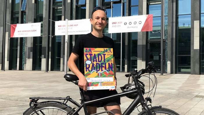 Der Stadttadeln-Botschafter der Universität Leipzig Daniel Obst steht mit seinem Fahrrad vor dem Paulinum und hält ein Werbeplakat für das Stadtradeln in der Hand.
