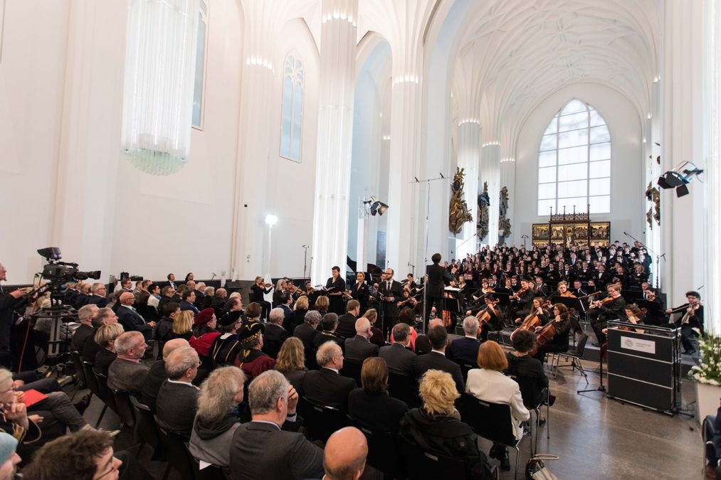 enlarge the image: Farbfoto: Blick auf den Altarbereich im Paulinum, wo Universitätsorchester und Chor musizieren. Davor stehen viele Stühle, auf denen festlich gekleidete Menschen sitzen.