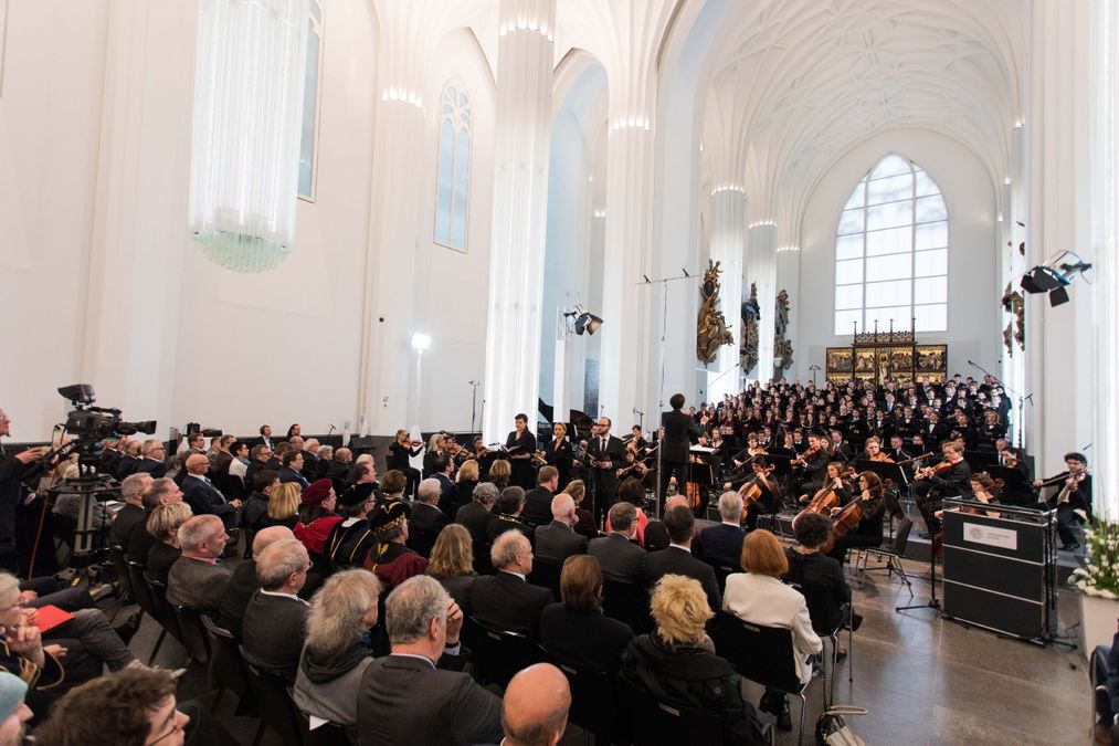 zur Vergrößerungsansicht des Bildes: Farbfoto: Blick auf den Altarbereich im Paulinum, wo Universitätsorchester und Chor musizieren. Davor stehen viele Stühle, auf denen festlich gekleidete Menschen sitzen.