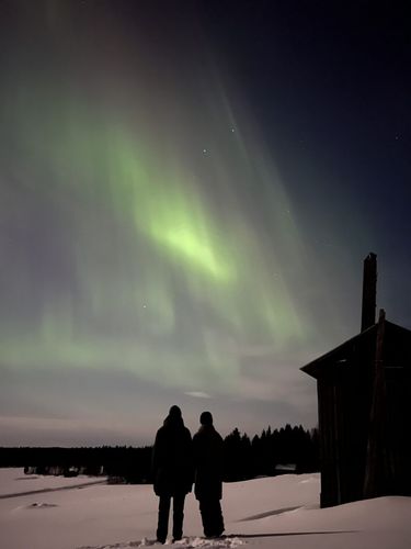 Zwei Personen blicken zu den Polarlichtern über verschneiter Landschaft.
