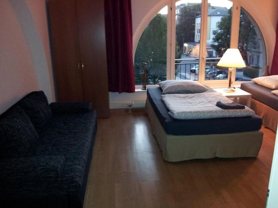 enlarge the image: Farbfoto: Innenaufnahme eines Wohnraums mit einem Sofa, einem Bett, einer Stehlampe und bodentiefen Fenster