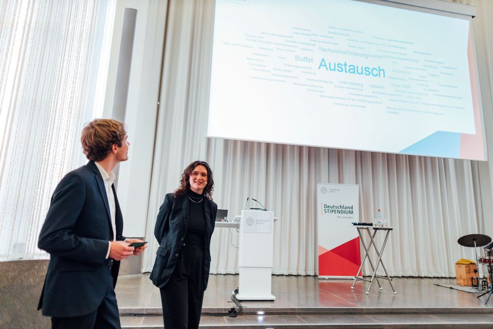 zur Vergrößerungsansicht des Bildes: die beiden Moderatoren der Veranstaltung stehen vor der Bühne, im Hintergrund ist die Leinwand mit Umfrageergebnissen