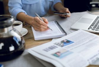 Schreibtisch mit Blocl, Laptop und Kaffeekanne, Frau schaut aufs Smartphone