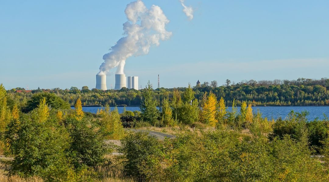 Farbige Naturaufnahme eines Sees, grüner Böschung, Im Hintergrund sind zwei Schornsteine eines Kraftwerks, die viel Dampf aussondern.