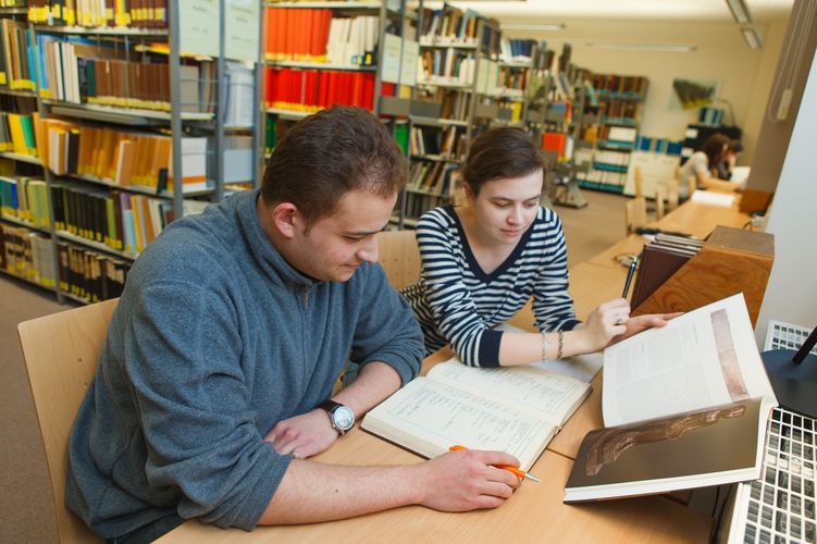Zwei Studierende sitzen beisammen in der Bibliothek und gehen Bücher durch, Foto: Christian Hüller