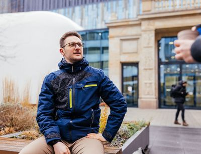Studienbotschafter Sebastian sitzt auf dem Campus und spricht mit einem Studenten, Foto: Christian Hüller