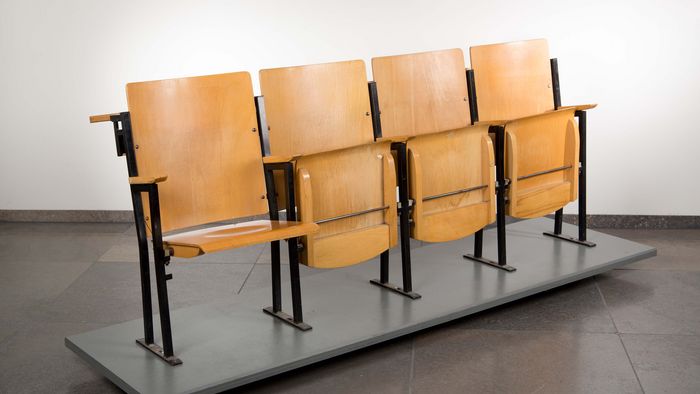 Das Foto zeigt eine Sitzreihe mit vier Stühlen aus einem der großen Hörsäle der Karl-Marx Universität aus dem Jahr 1978. Die Hörsaalbank, bestehend aus hellbraunem Birkenholz und schwarzem Metallgerüst, ist frontal, aber leicht schräg vor grauem Hintergrund dargestellt. Die Sitzfläche des Stuhls ganz links ist nach unten geklappt, die der übrigen drei ist nicht ausgeklappt. 