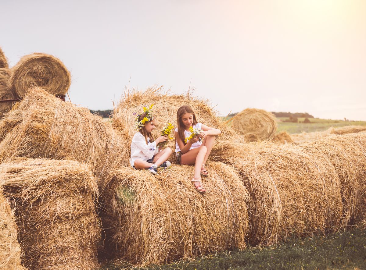 Man sieht zwei Mädchen mit Blumen in den Haaren auf Heuballen sitzen. Im Hintergrund steht die Sonne tief.