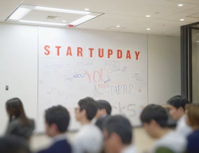 Im Vordergrund sind sitzende Zuhörer, im Hintergrund eine weiße Wand mit Aufschrift "Startupday" zu sehen