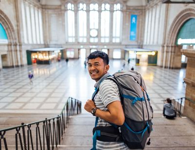 Colour photo: A person with a rucksack smiles at the camera. The entrance hall of Leipzig railway station is in the background.