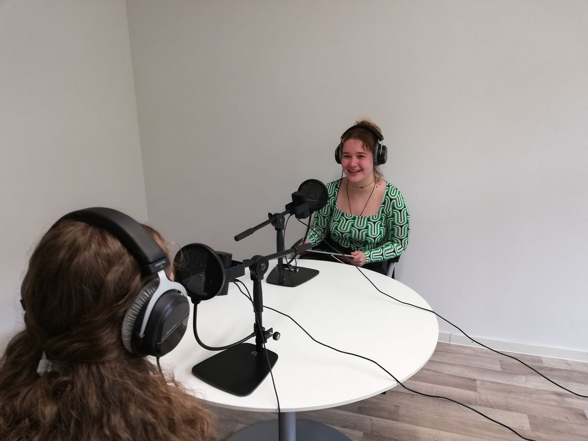 zur Vergrößerungsansicht des Bildes: Zwei Studierende sitzen sich an einem Tisch gegenüber und nutzen Mikrofontechnik zur Aufnahme des Podcasts.