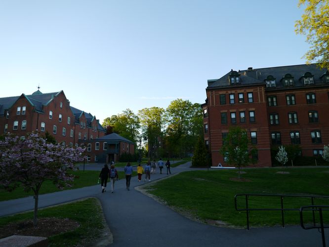 Abgebildet ist eine Wegkreuzung auf dem Campusgelände, welche sich inmitten einer grünen Wiese erstreckt. Man sieht einige Studierende, die auf dem Weg spazieren. Im Hintergrund sind rote Backsteinhäuser und blühende Bäume zu erkennen.