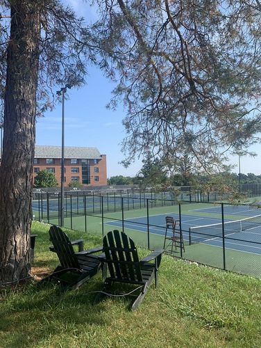 Blaue Tennisplätze sind im Hintergrund des Bildes zu sehen. Das Foto ist von einer Anhöhe aufgenommen. Auf dieser Anhöhe steht ein Baum und zwei Gartenstühle.