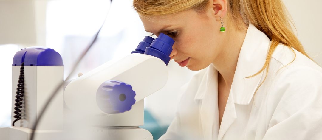 Foto: eine Frau sitzt im Arztmantel am Mikroskop