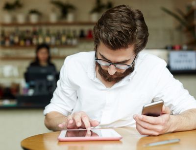 Person sitzt in einem Café, hält ein Handy in der einen Hand, während die andere Hand auf ein Tablet tippt.