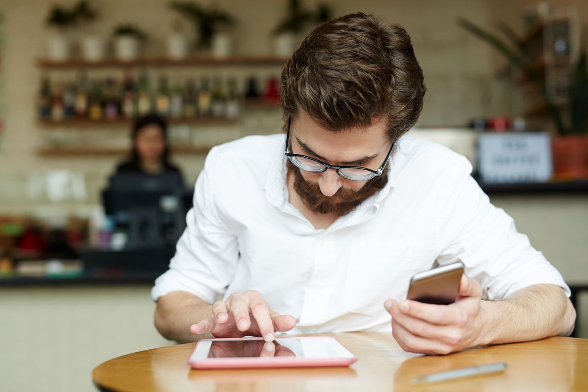  Person sitzt einem Café, mit einem Handy in der einen Hand, während die andere auf einem Tablet tippt.