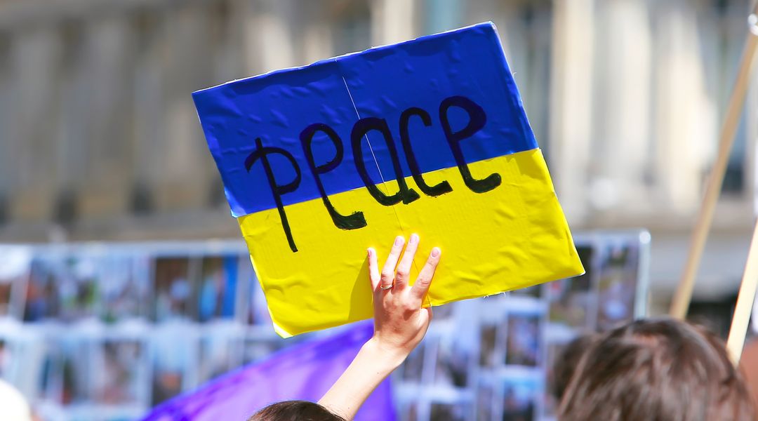 Farbfoto: Demonstrationszug, eine Frau hält ein handgemaltes Pappschild in den Farben der ukrainischen Flagge in die Höhe, auf dem mit schwarzen Buchstaben das Wort "peace" (Frieden) steht.