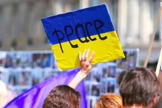 Farbfoto: Demonstrationszug, eine Frau hält ein handgemaltes Pappschild in den Farben der ukrainischen Flagge in die Höhe, auf dem mit schwarzen Buchstaben das Wort "peace" (Frieden) steht.