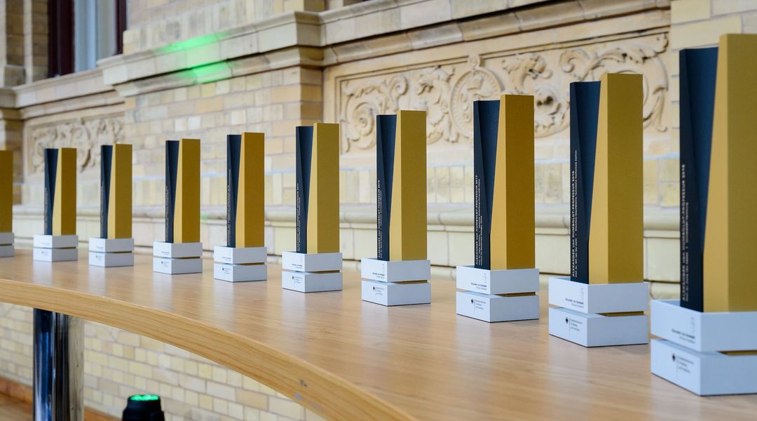 In einer Reihe stehen die Preise, schwarz gelbe Trophäen, für die elf angehenden Humboldt-Profesor:innen auf einem langen Tisch.