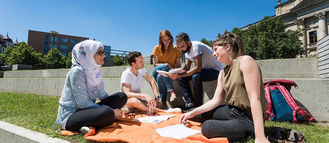Internationale Studierende sitzen auf einer Picknickdecke in der Sonne vor dem Campus Beethovenstraße 