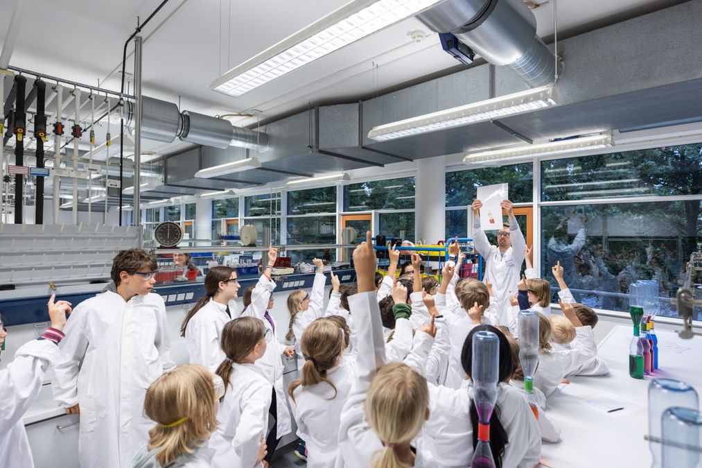 zur Vergrößerungsansicht des Bildes: Labor mit einem Mann in weißem Kittel um ihn herum stehen circa zwanzig Kinder, ebenfalls in weißen Kitteln. Einige Kinder melden sich.