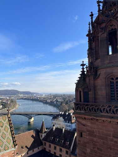 Das Foto wurde vom Basler Münster aus geschossen und blickt auf den Fluss Rhein. Am linken und rechten Bildrand sieht sieht man die Dächer von Basel vor blauem Himmel im Hintergrund.