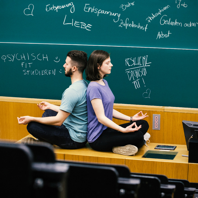 Zwei Studierende sitzen im Vorlesungssaal und meditieren