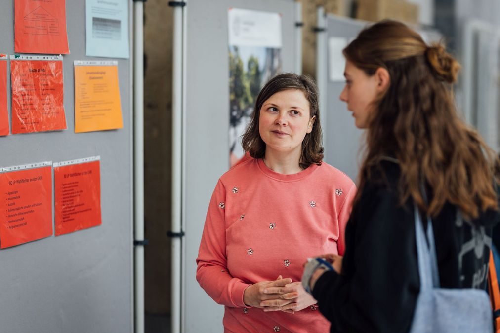 zur Vergrößerungsansicht des Bildes: Foto: Eine Mitarbeiterin der Fakultät für Geschichte, Kunst- und Regionalwissenschaften ist am Infostand im Gespräch mit einer Besucherin.