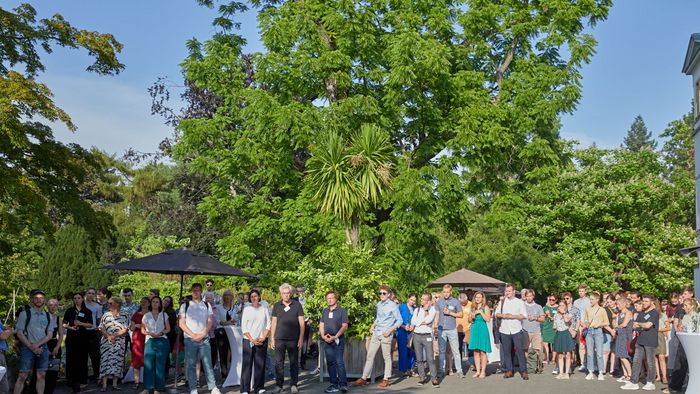 Förderer und Stipendiaten stehen an einem sonnigen Tag, auf einem freien Platz, vor einem großen Baum im Botnischen Garten und hören der Rede der Rektorin, Frau Prof. Obergfell zu.