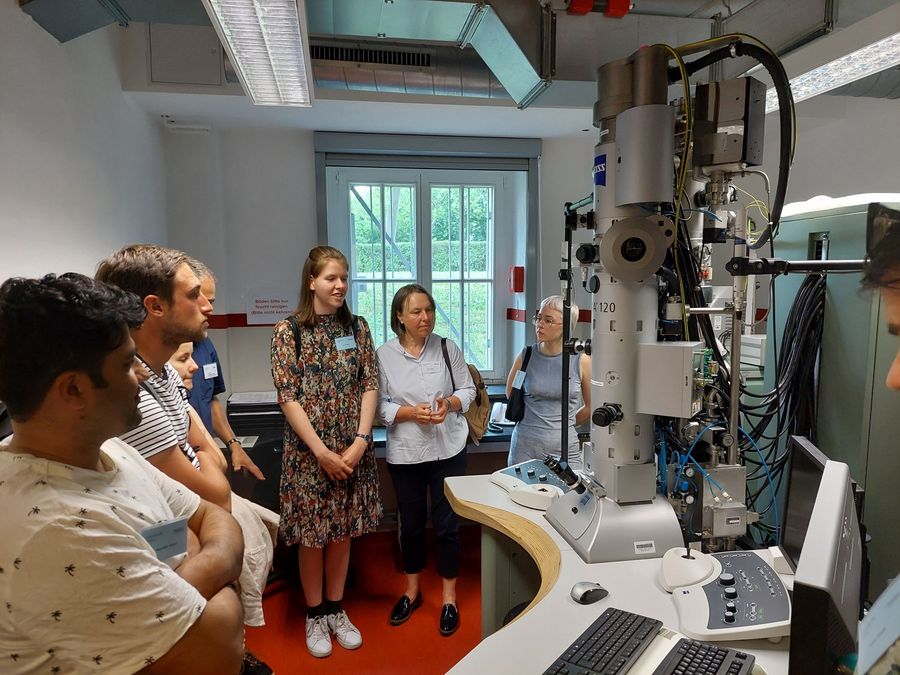 zur Vergrößerungsansicht des Bildes: Farbfoto: Förder:innen und Stipendiat:innen stehen in einem kleine Raum mit einem sehr großen Mikroskop