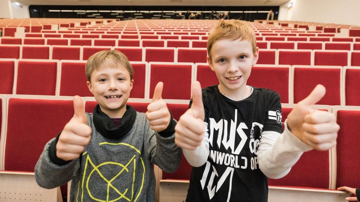 zur Vergrößerungsansicht des Bildes: Frontalaufnahme von zwei Jungen im Hörsaal, die in die Kamera lächeln und beide Dauemn hoch zeigen.