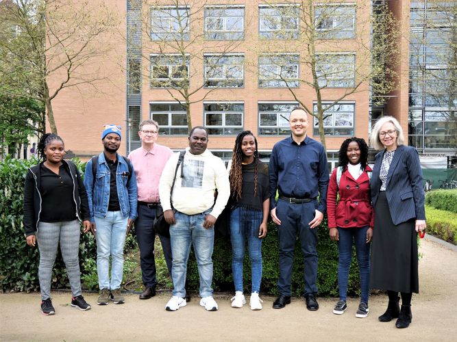 Gruppenfoto der fünf mosambikanischen Austauschstudierenden mit ihren drei Leipziger Betreuer:innen im Freien vor einer grünen Hecke. Im Hintergrund ist ein Teil der rötlichen Fassade des Geisteswissenschaftlichen Zentrums der Universität Leipozig zu sehen.