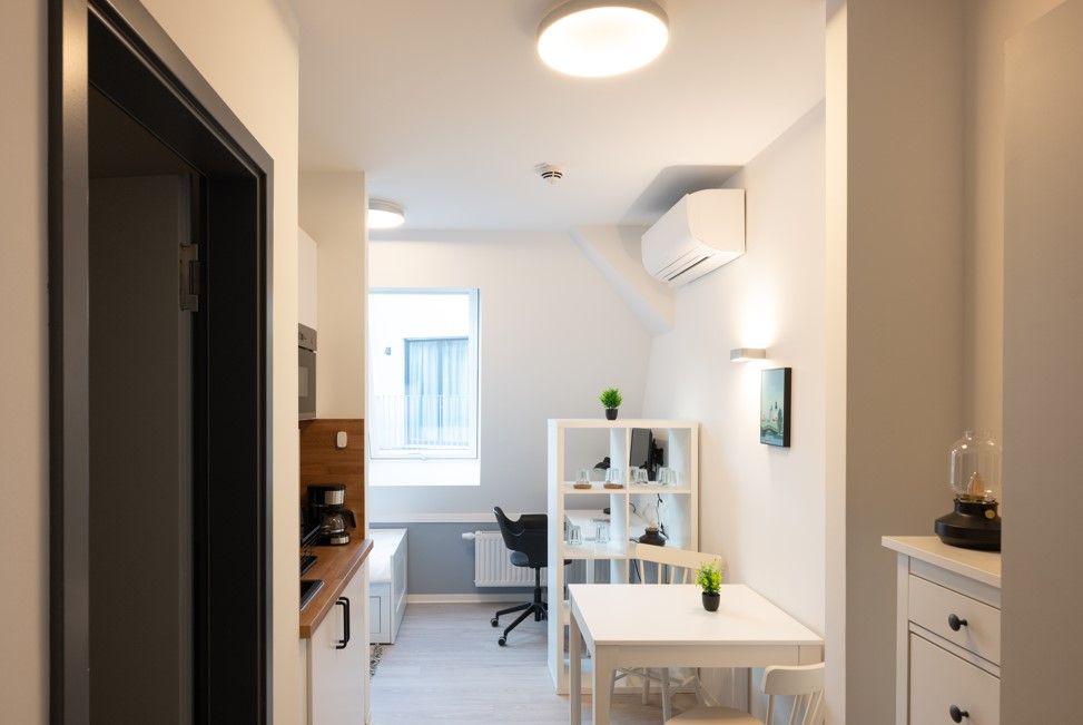 enlarge the image: View from the entrance area of a cozy apartment, showing a dining table with two chairs, a room divider with a workspace behind it, a large window, air conditioning, and a small kitchenette. Photo: Thomas Biermann