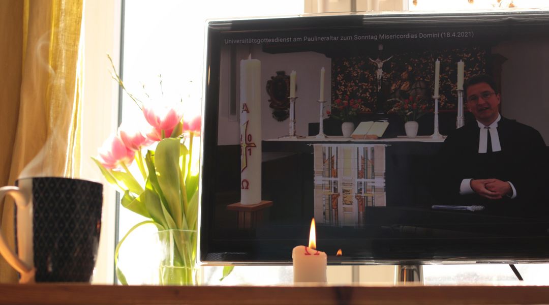 Auf einem Schreibtisch steht ein Bildschirm, worauf ein Video des Universitätsgottesdienstes läuft. Daneben steht ein Straß Tulpen, eine Kerze und eine dampfende Kaffeetasse.