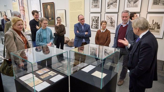 Förderer und Stipendiaten stehen um eine Glasvitrine in der Galerie im Neuen Augusteum und hören dem Kustos, der vor den Gästen steht, zu