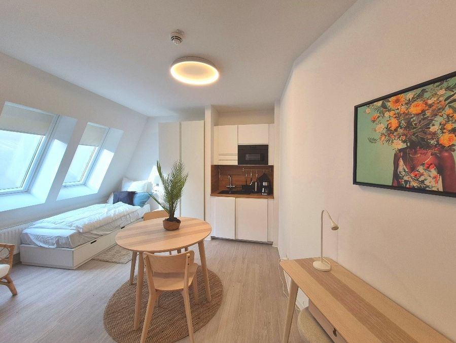 enlarge the image: Small apartment with a single bed, wardrobe, a round table with two chairs in the center of the room, a small kitchenette, a colorful wall art, and a workspace. Photo: Thomas Biermann