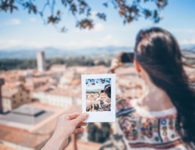 Die linke Hand einer Person hält vor einen verschwommenen Hintergrund ein Polaroid-Foto auf dem eine Frau abgebildet ist. Die Frau blickt auf eine Stadt und macht ein Foto.